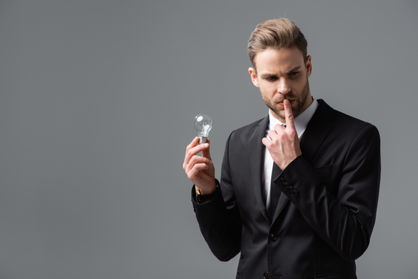 hombre de negocios pensativo tocando los labios mientras sostiene la bombilla aislada en gris - Foto, imagen