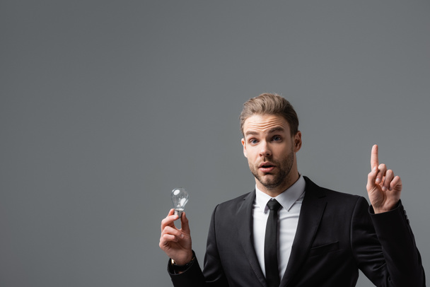 возбужденный бизнесмен показывает идею жест, держа лампочку изолированы на серый - Фото, изображение