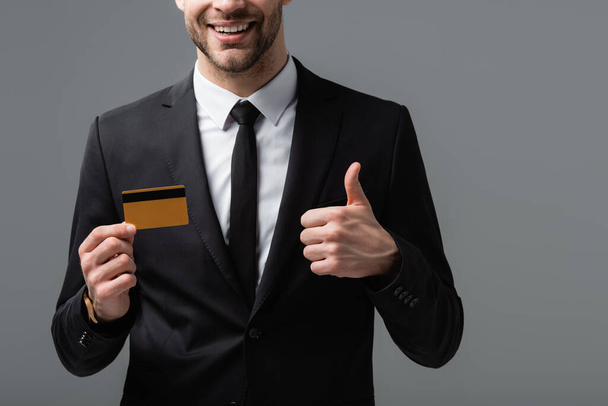 vista parcial del gerente alegre mostrando el pulgar hacia arriba y la tarjeta de crédito aislada en gris - Foto, Imagen