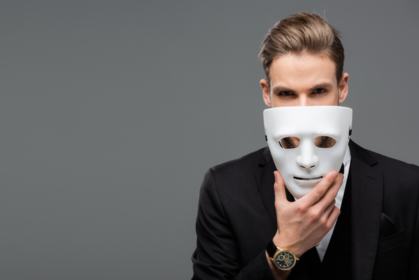 hombre de negocios sosteniendo la máscara cerca de la cara mientras mira la cámara aislada en gris - Foto, Imagen