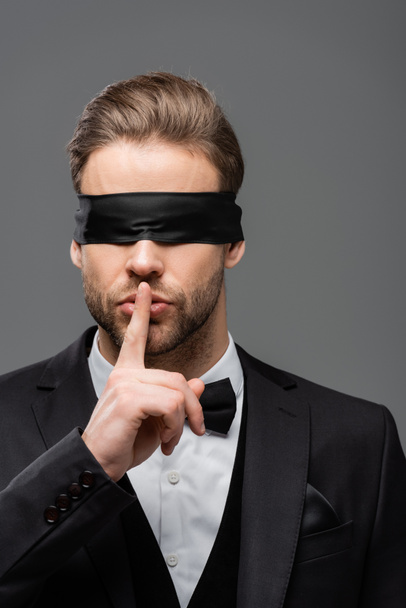 junger Geschäftsmann mit verbundenen Augen zeigt Schweigezeichen isoliert auf grau - Foto, Bild