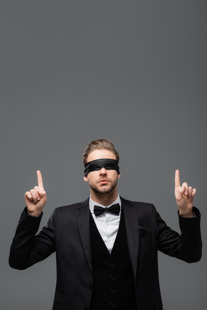 young, blindfolded businessman pointing up with fingers isolated on grey - Photo, Image