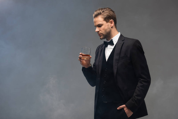 junger Geschäftsmann mit Whiskey in der Hand, während er mit der Hand in der Tasche vor grauem Hintergrund mit Rauch steht - Foto, Bild