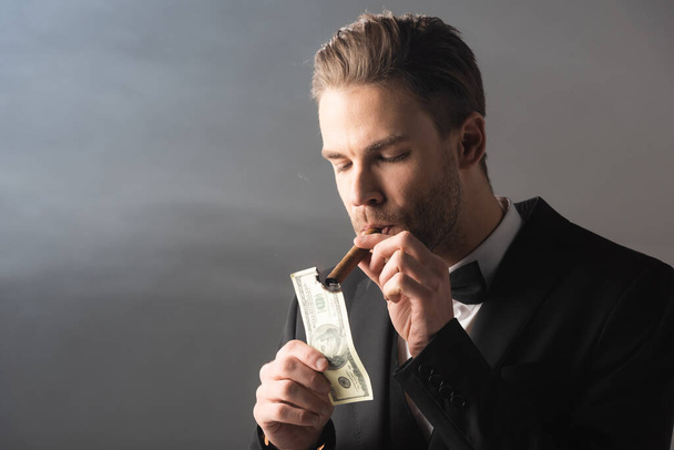 молодой бизнесмен закуривает сигару со стодолларовой купюрой на сером фоне с дымом - Фото, изображение