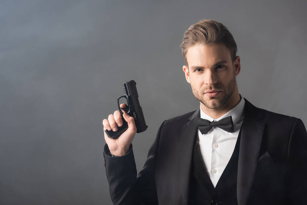 homem de negócios elegante olhando para a câmera enquanto segurava a arma no fundo cinza com fumaça - Foto, Imagem