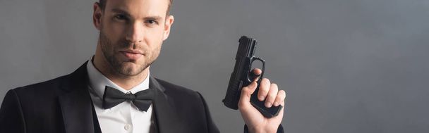 serious businessman looking at camera while holding gun on grey background with smoke, banner - Photo, Image