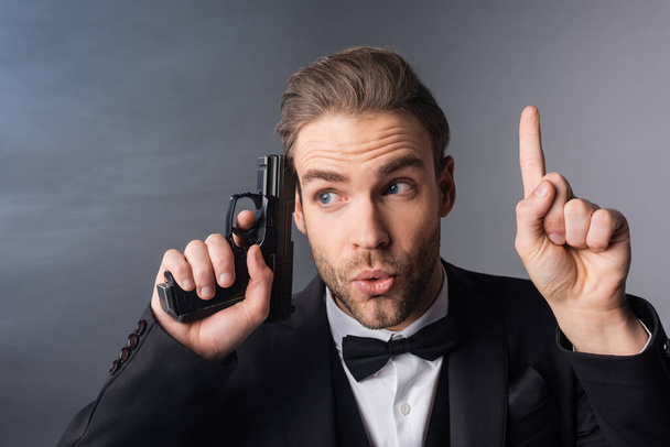 uomo d'affari eccitato mostrando gesto idea mentre tiene arma vicino alla testa su sfondo grigio con fumo - Foto, immagini