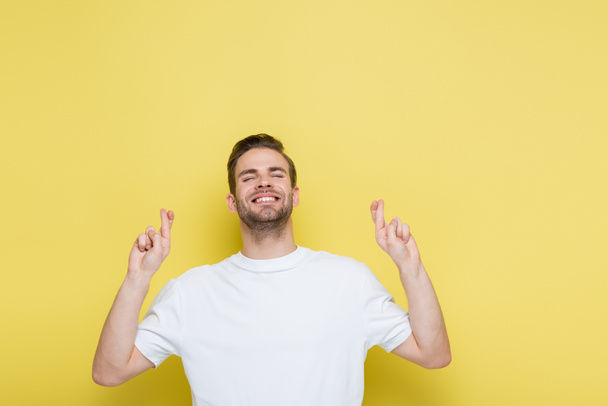 vrolijke man met gesloten ogen staande met gekruiste vingers op geel - Foto, afbeelding