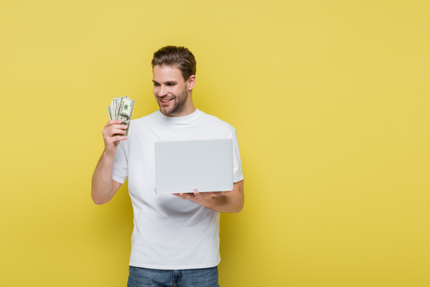 Bilgisayarı sarıda tutarken dolarlara bakan memnun bir adam. - Fotoğraf, Görsel