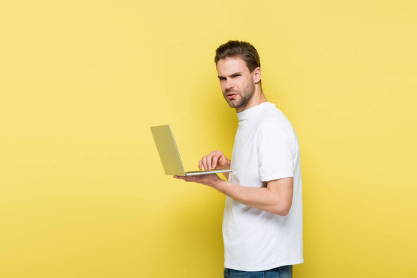 Dizüstü bilgisayarı sarıda yazarken kameraya bakan hoşnutsuz adam. - Fotoğraf, Görsel