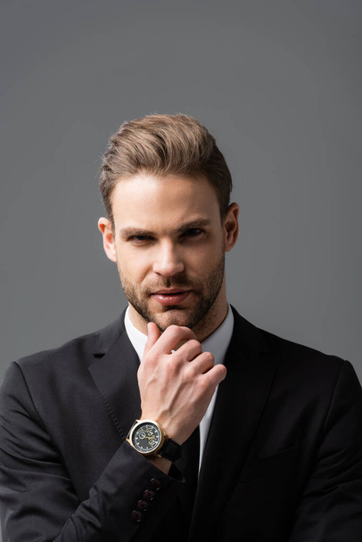 young, confident businessman looking at camera while touching chin isolated on grey - Photo, Image