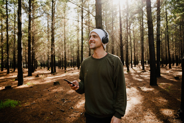 Caucasian male holding smartphone playing relaxing music wearing headphones having a good time in luscious woodlands  - Zdjęcie, obraz