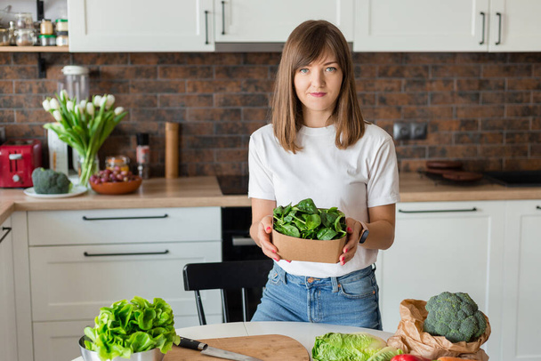 Woman in white t-shirt holding a green spinach in recycling ecology packing in kitchen at home. Raw food diet and healthy eating concept. Baby food - Photo, Image