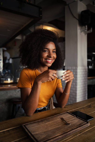 Good looking African American female sitting contently gazing while holding coffee mug sitting in trendy tearoom. - Foto, afbeelding