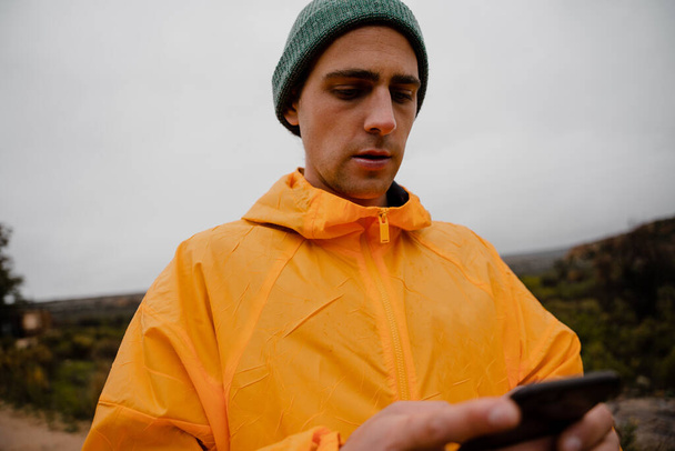 Çekici genç erkek yürüyüşçü bulutlu bir havada dik dağ yürüyüşü yolunda akıllı telefon kullanarak yön bulmaya odaklanıyor. - Fotoğraf, Görsel