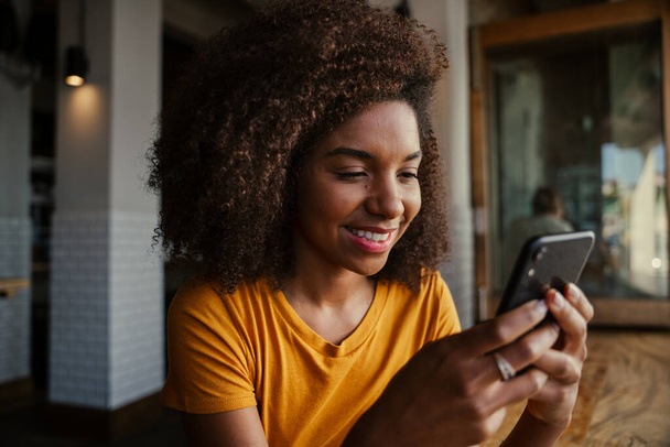 Красивая улыбающаяся этническая клиентка-подросток получает сообщение от парня на смартфоне в модном кафе  - Фото, изображение