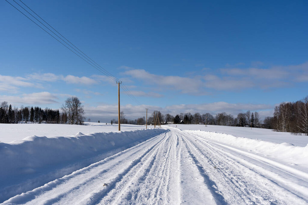 красивый солнечный зимний день в латвийской сельской местности с голубым небом и белыми облаками, где посреди поля лежит покрытая снегом дорога, на которой следы автомобильных шин остаются в снегу - Фото, изображение