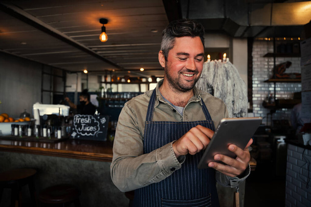 Smiling white male coffee shop owner scrolling on tablet in fashionable coffee shop. - Photo, Image