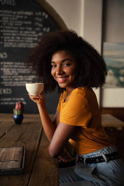 Вишукана етнічна жінка, посміхаючись на камеру, тримає гарячу каву, сидячи в модному кафе
. - Фото, зображення