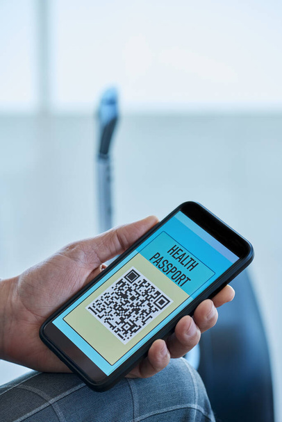 ein junger kaukasischer Geschäftsmann in einem eleganten grauen Anzug sitzt in der Wartehalle eines Flughafens und hat einen simulierten elektronischen Gesundheitsausweis auf dem Bildschirm seines Smartphones - Foto, Bild