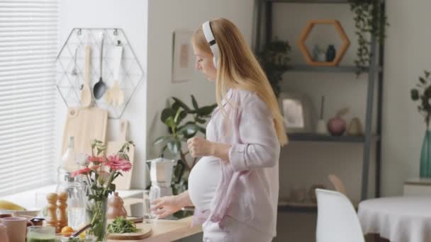 Iloinen raskaana oleva nainen langattomissa kuulokkeissa tanssii musiikkia ja juo lasillista vettä keittiössä kotona - Materiaali, video