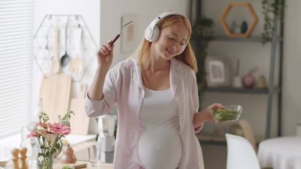 Vidám terhes nő vezeték nélküli fejhallgatóval kezében tál saláta és villa, énekel és táncol a zene a konyhában otthon - Felvétel, videó