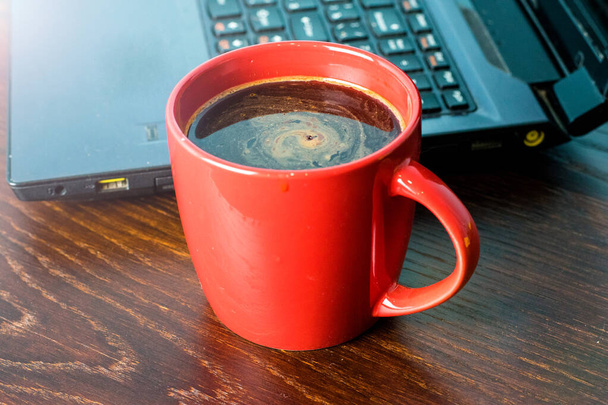 Egy csésze kávé és laptop a háttérben egy barna fa asztalon. A távmunka vagy képzés fogalma, a munkanap kezdete az irodában. - Fotó, kép