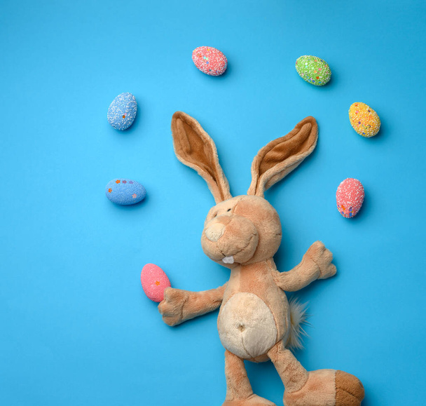 dekoracyjne pisanki i miękka pluszowa zabawka królicza z długimi uszami na niebieskim tle, jajka lecą na górze - Zdjęcie, obraz