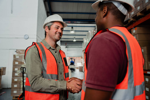 Lächelnder kaukasischer Fabrikmann beim Händeschütteln mit einem Mitarbeiter, der im Lager Geschäfte macht - Foto, Bild