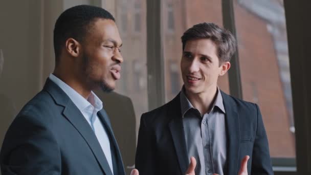 Kaksi monietnistä monirotuista miestä muodollisissa puvuissa pari liikemiestä afrikkalainen amerikkalainen mies ja kaukasialainen työntekijä seisovat lähellä toimiston ikkunaa puhumassa väittävät suunnittelevansa nauramista aktiivisesti - Materiaali, video