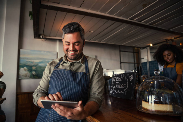 Άνδρας σερβιτόρος περιήγηση σε ψηφιακή tablet κύλιση μέσω μενού στέκεται ενάντια στον πάγκο σε funky cafe  - Φωτογραφία, εικόνα