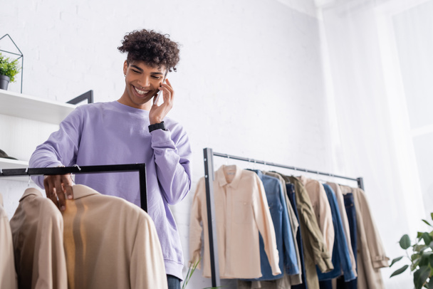 Sonriente afroamericano propietario de sala de exposición hablando en el teléfono inteligente cerca de la ropa en primer plano borrosa  - Foto, imagen