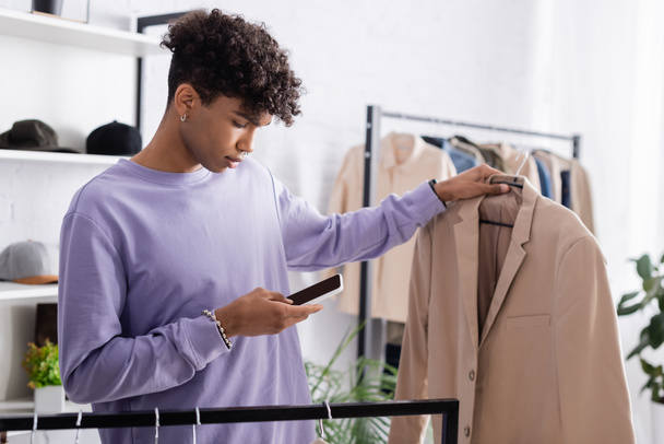 African american showroom owner using smartphone and holding jacket on hanger on blurred background  - Photo, Image