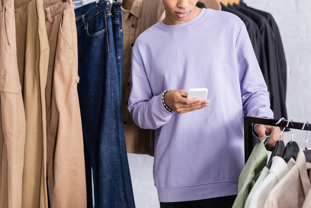 Vue recadrée du propriétaire afro-américain du showroom en utilisant un smartphone près des vêtements  - Photo, image