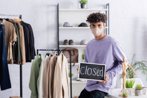 African american owner of showroom in protective mask holding chalkboard with closed lettering during quarantine  - Photo, Image