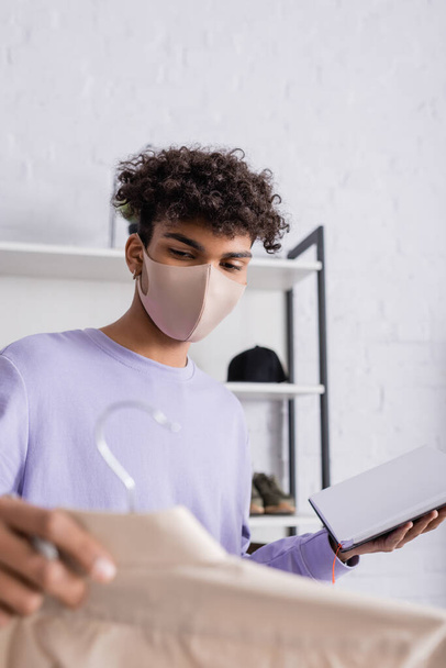 African american owner of showroom in protective mask holding notebook and shirt on hanger on blurred foreground  - Photo, Image