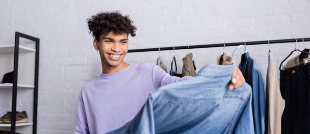 Proprietario di showroom afroamericano sorridente che tiene la camicia di denim in primo piano sfocata, banner  - Foto, immagini