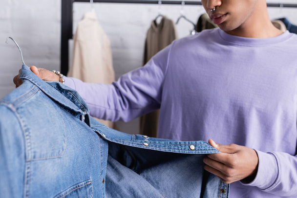 Ausgeschnittene Ansicht eines Jeanshemdes in der Hand eines afrikanisch-amerikanischen Showroom-Besitzers auf verschwommenem Hintergrund  - Foto, Bild