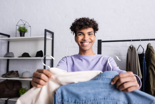 Afro-Amerikaanse eigenaar van showroom glimlachen op camera en het houden van shirts op hangers op wazig voorgrond  - Foto, afbeelding