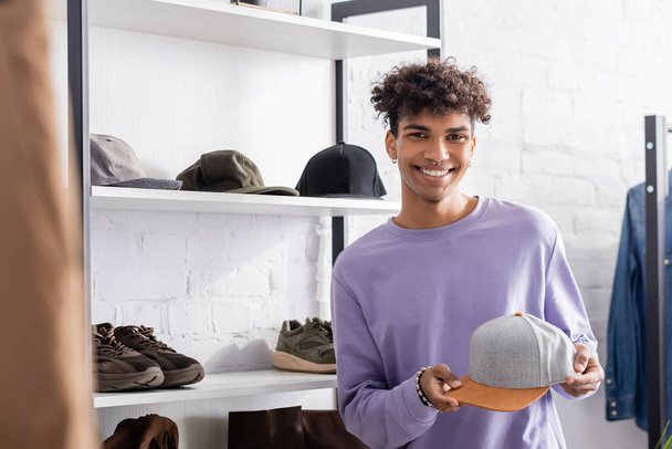 Smiling african american owner of showroom looking at camera while holding cap  - Photo, Image