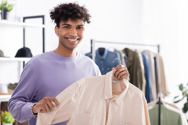 Χαμογελώντας αφροαμερικανός ιδιοκτήτης του showroom κρατώντας πουκάμισο στην κρεμάστρα και χαμογελώντας στην κάμερα  - Φωτογραφία, εικόνα