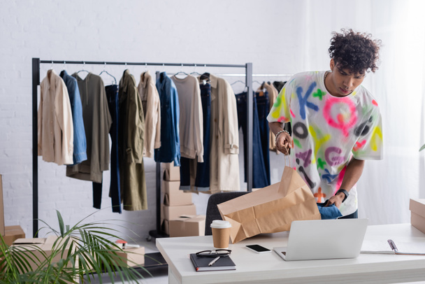 Propietario afroamericano de showroom poniendo ropa en bolsa de compras cerca de dispositivos y café para llevar  - Foto, imagen