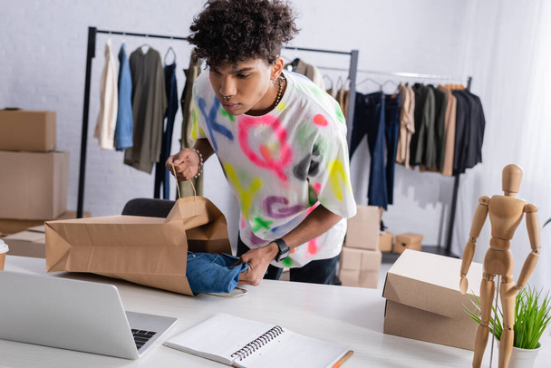 Africano americano proprietário de showroom segurando saco de compras e camisa perto de laptop e caixa  - Foto, Imagem