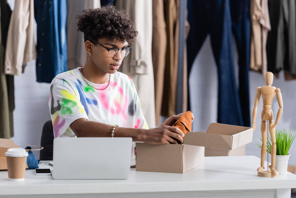 Νεαρός Αφροαμερικάνος επιχειρηματίας που βάζει ρούχα σε χαρτοκιβώτιο κοντά σε laptop και καφέ για να πάει  - Φωτογραφία, εικόνα