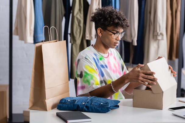 Proprietario di piccola impresa afroamericana che tiene la scatola vicino alla borsa della spesa e ai vestiti nello showroom  - Foto, immagini