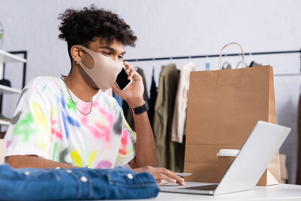 Proprietario afroamericano di showroom in maschera protettiva che parla su smartphone e utilizza laptop vicino alla shopping bag  - Foto, immagini