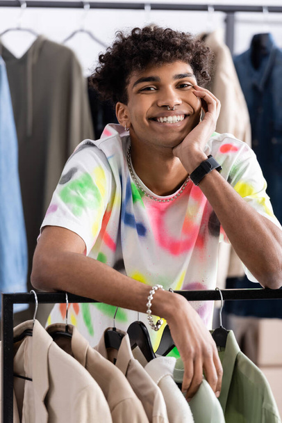 Χαμογελώντας αφροαμερικανός ιδιοκτήτης εκθεσιακού χώρου κοιτάζοντας κάμερα κοντά σε ρούχα  - Φωτογραφία, εικόνα