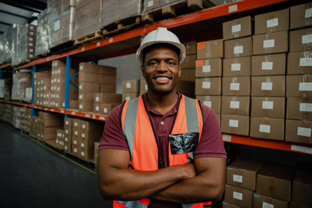 Primer plano del trabajador masculino con brazos cruzados sonriendo mientras está de pie junto a los paquetes en la tienda de la fábrica  - Foto, Imagen