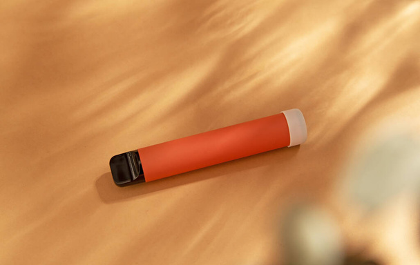 оранжевая палочка - одноразовое вапирующее устройство, электронная сигарета на бежевом фоне. Плоская кладка, вид сверху, минимальный дизайн с тенями солнечного света - Фото, изображение