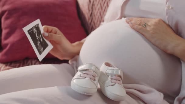 Zbliżenie widok oczekującej matki siedzącej na kanapie z uroczymi bucikami na kolanach, trzymającej obraz USG płodu i pocierającej brzuch w ciąży czekając na dziecko - Materiał filmowy, wideo